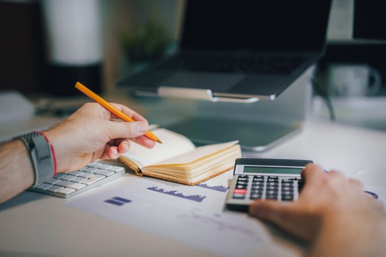 A person working at a laptop with receipts, notebooks, and a calculator on a kitchen table.
